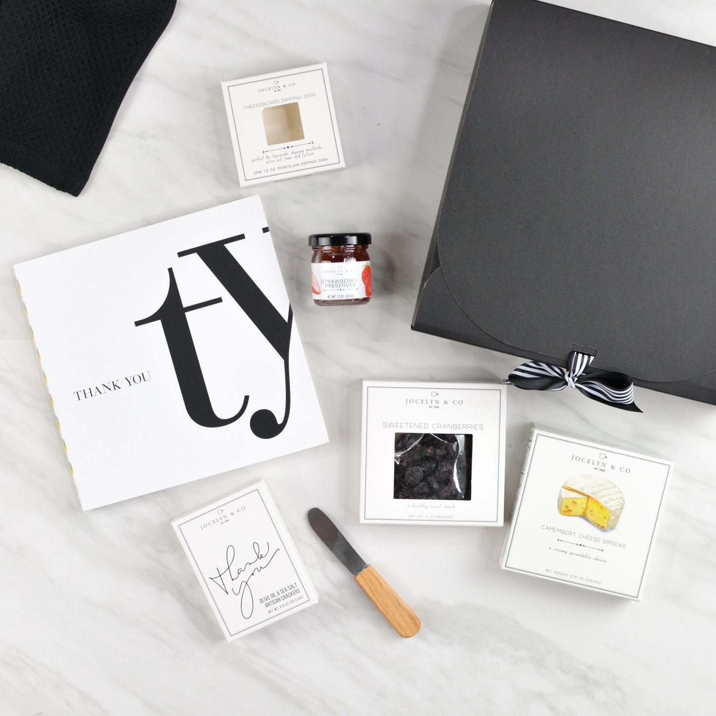 Black thank-you gift box with gratitude book, gourmet crackers, cheese, jam, and wooden spreader arranged neatly on a white surface