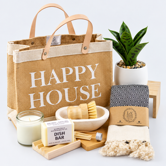 A curated bath and home gift set arranged on a white background, featuring a large tan burlap tote bag with 'HAPPY HOUSE' printed in white, a small green plant in a white pot, a candle, a dish bar, wooden utensils, a patterned towel, a scrubber, and a fluffy cream towel.