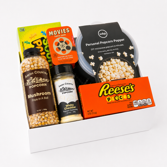 A curated popcorn gift set arranged in a white box, featuring a jar of Amish Country Popcorn Mushroom Pops in A Ball, a jar of Amish Country Popcorn White Cheddar Pops, a black wrap Personal Microwave Popcorn Popper, a box of Reese's Pieces, and a green and orange 'Movies for the Home' box.