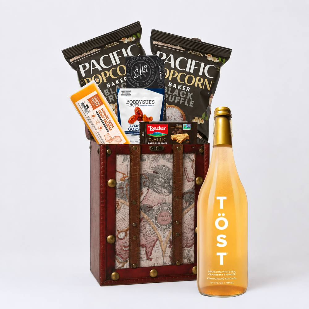 A curated gift basket or snack hamper set on a light gray studio background, featuring a wooden crate with vintage map artwork and leather straps, filled with snacks including bags of Pacific Popcorn, a box of Bobby's Cheese, and a small package of Tucker Classic Cigarettes, alongside a large bottle of Töst rosé wine.